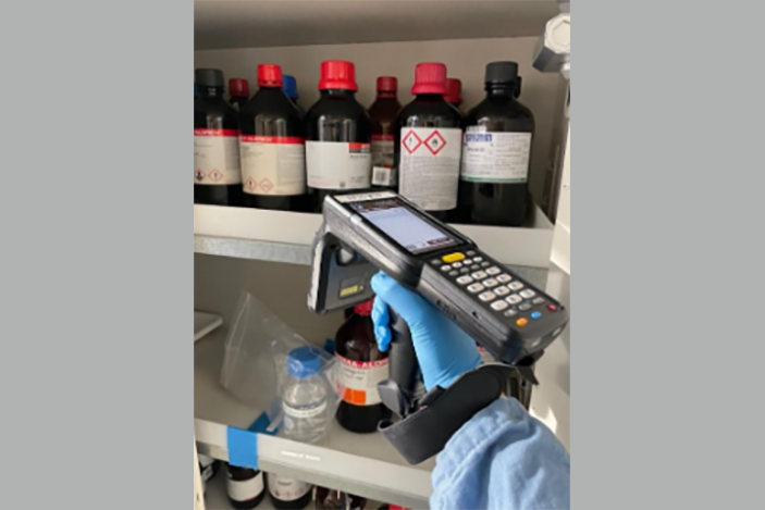 Closeup up of a person's gloved hand holding portable inventory device with bottles of chemicals in background.