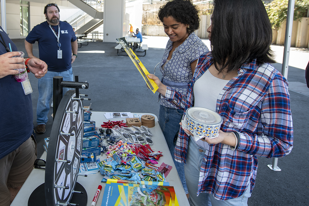 Two people at outside table with prize wheel and prizes on table