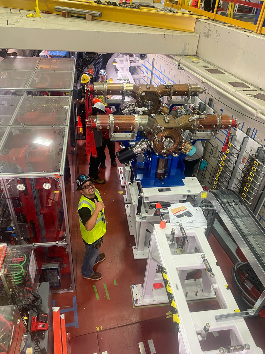 A person wearing a safety hat, goggles, and vest holding a thumbs up with equipment being installed in the background.