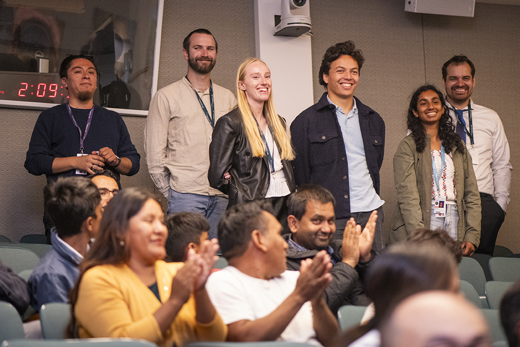 Six people standing in row at back of auditorium while people sitting in foreground applaud