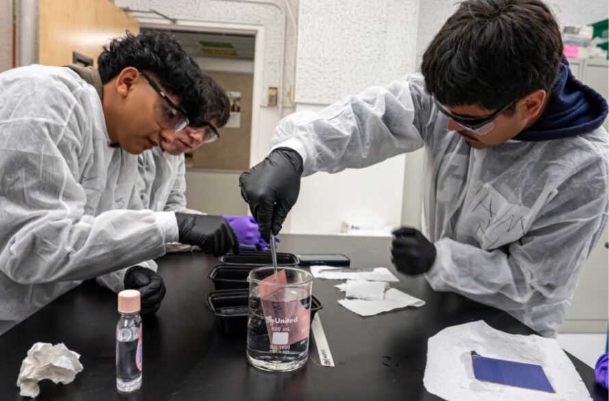Two young people in lab suits, safety glasses, and gloves in a lab