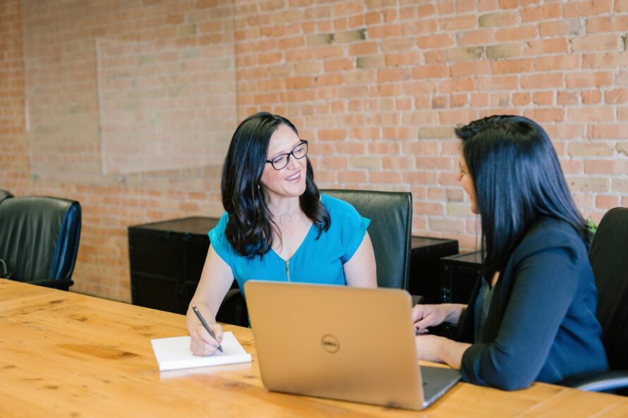 Two persons seated in front of an open laptop computer engaged in conversation.