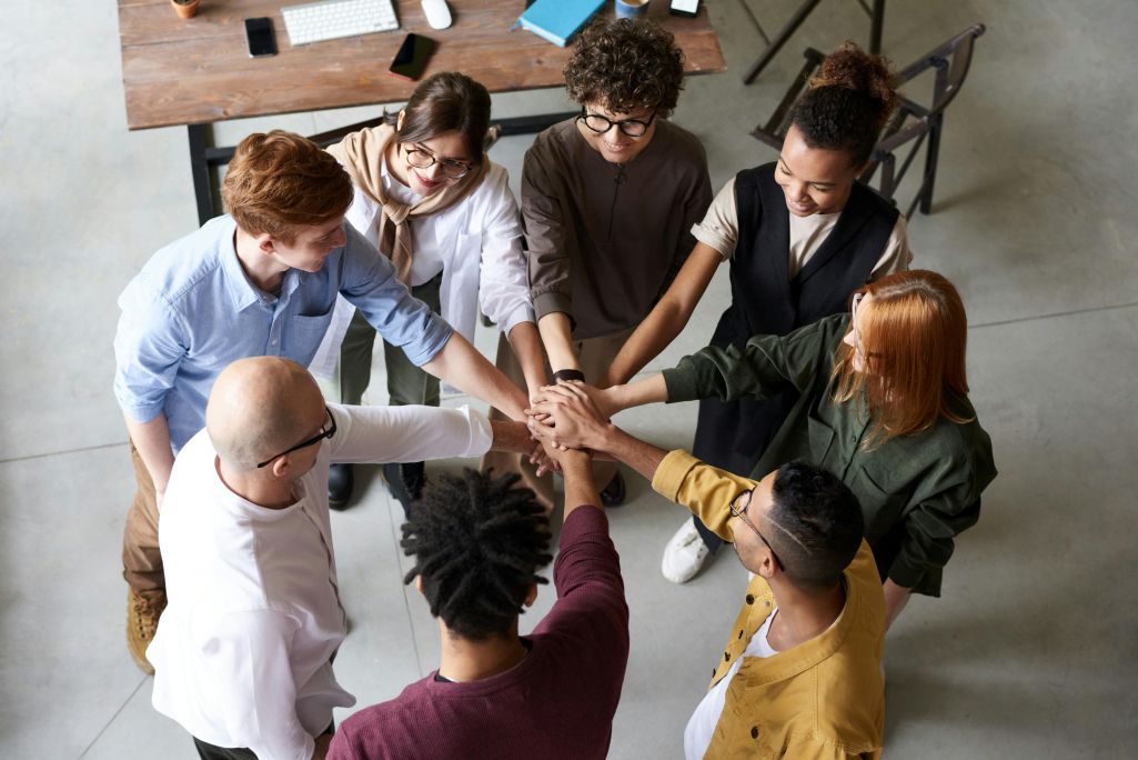 Group of persons standing in circle with arms outstretched with hands overlapping in center of circle.