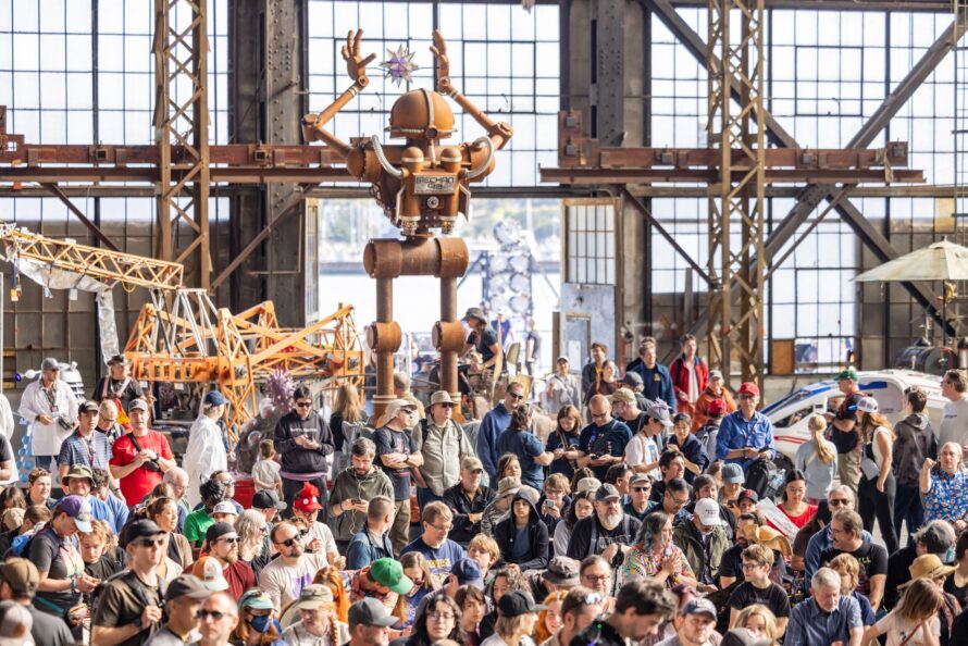 A crowd of persons standing in large meeting space with towering human-like robot standing in background.