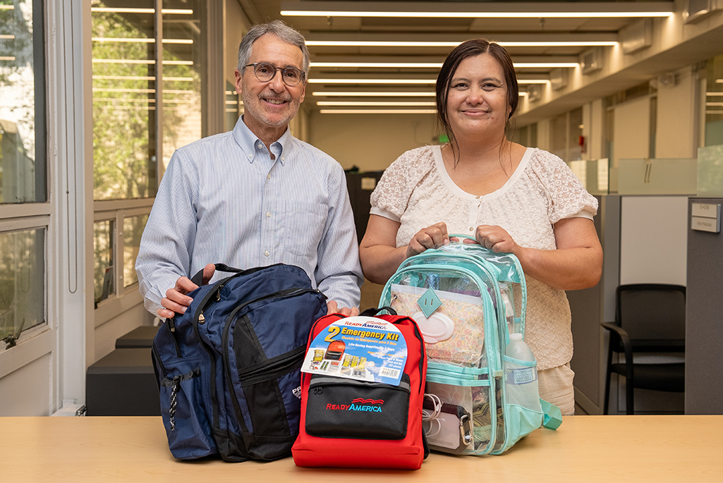 Two people standing behind a table with three backpacks in front of them.