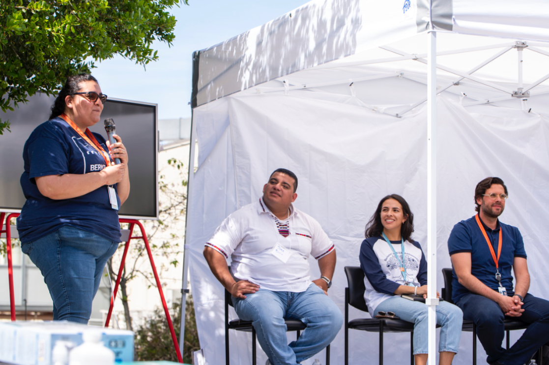 Person standing and talking into microphone with three other persons seated beside a white canopy tent listening.