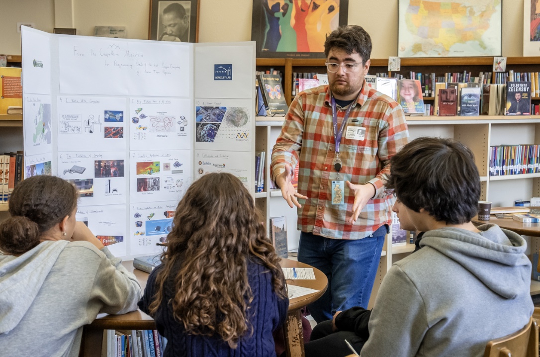 Person with dark hair and light glasses standing in front of poster gesturing toward three seated people looking on.