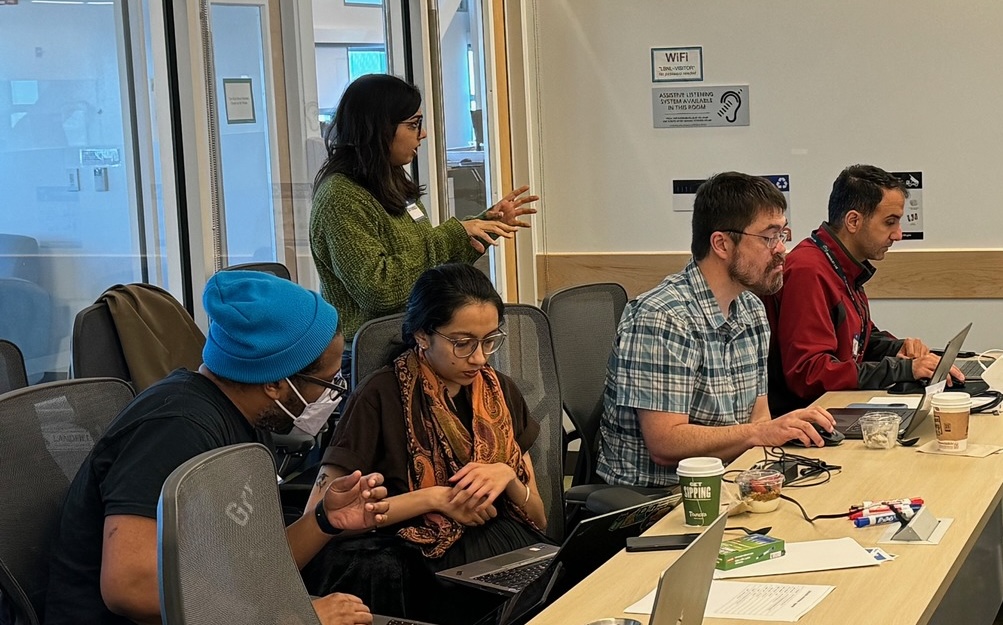 Several persons seated at table with person behind them standing and talking.
