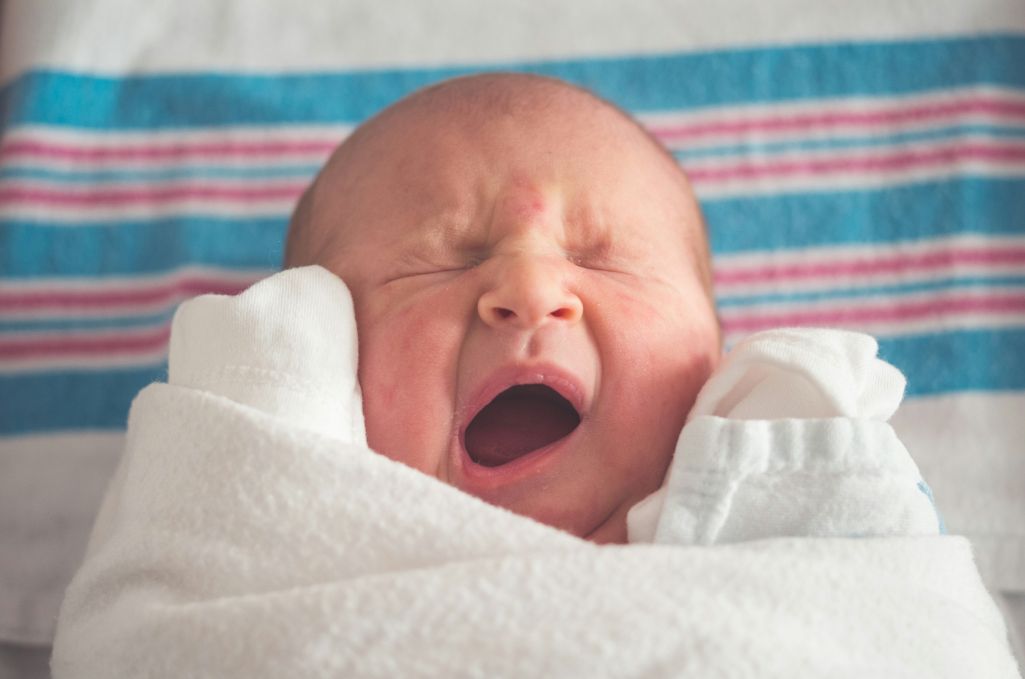 Photo of infant swaddled in white blanket, yawning, and resting on blue and red-striped background.
