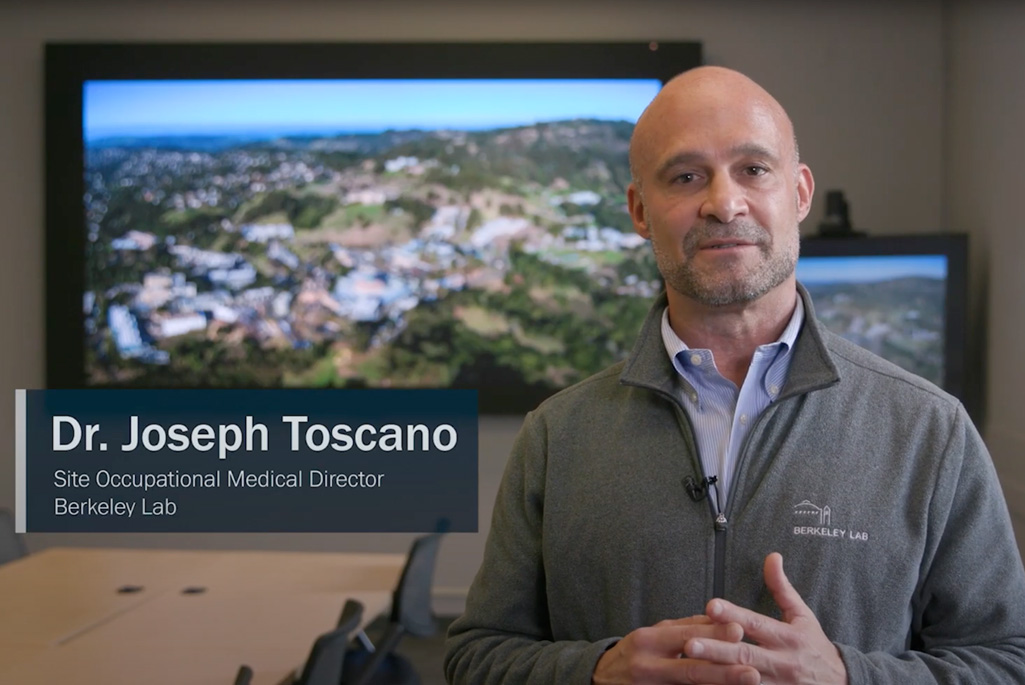 Person wearing a gray jacket with the Berkeley Lab logo speaking in a conference room. Text on screen says, "Dr. Joseph Toscano, Site Occupational Medical Director, Berkeley Lab."