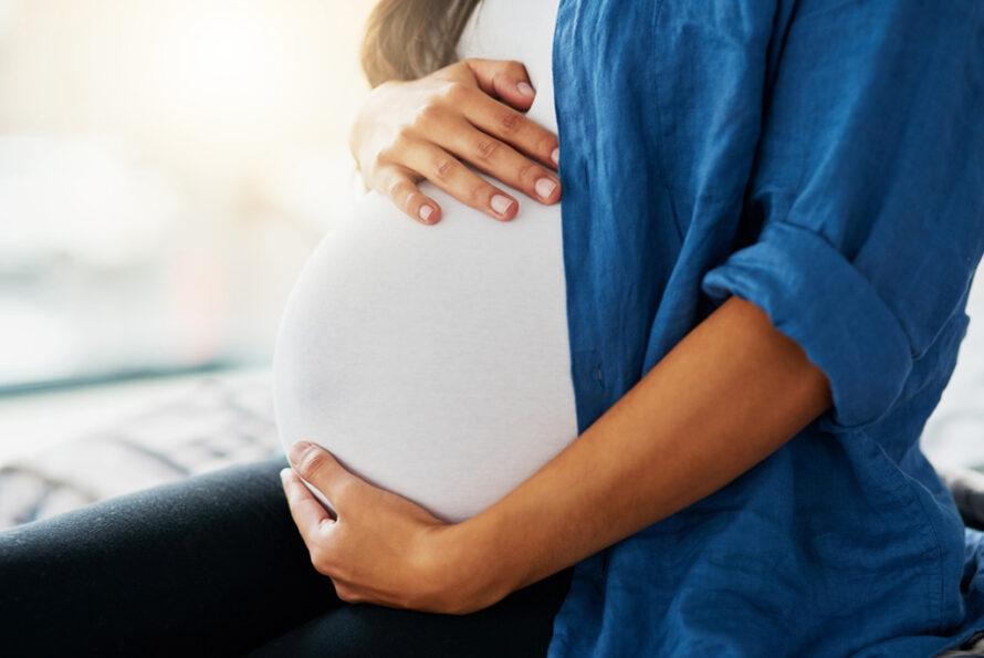 Close-up image of a pregnant person wearing a white top and blue jacket holding their belly.