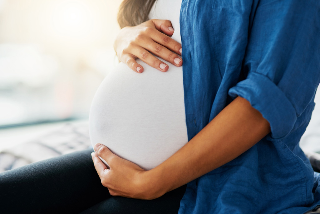 Close-up image of a pregnant person wearing a white top and blue jacket holding their belly.