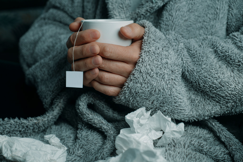 A sick person at home, wearing a fluffy gray house robe, surrounded by used tissues, warming up with a cup of hot herbal tea.