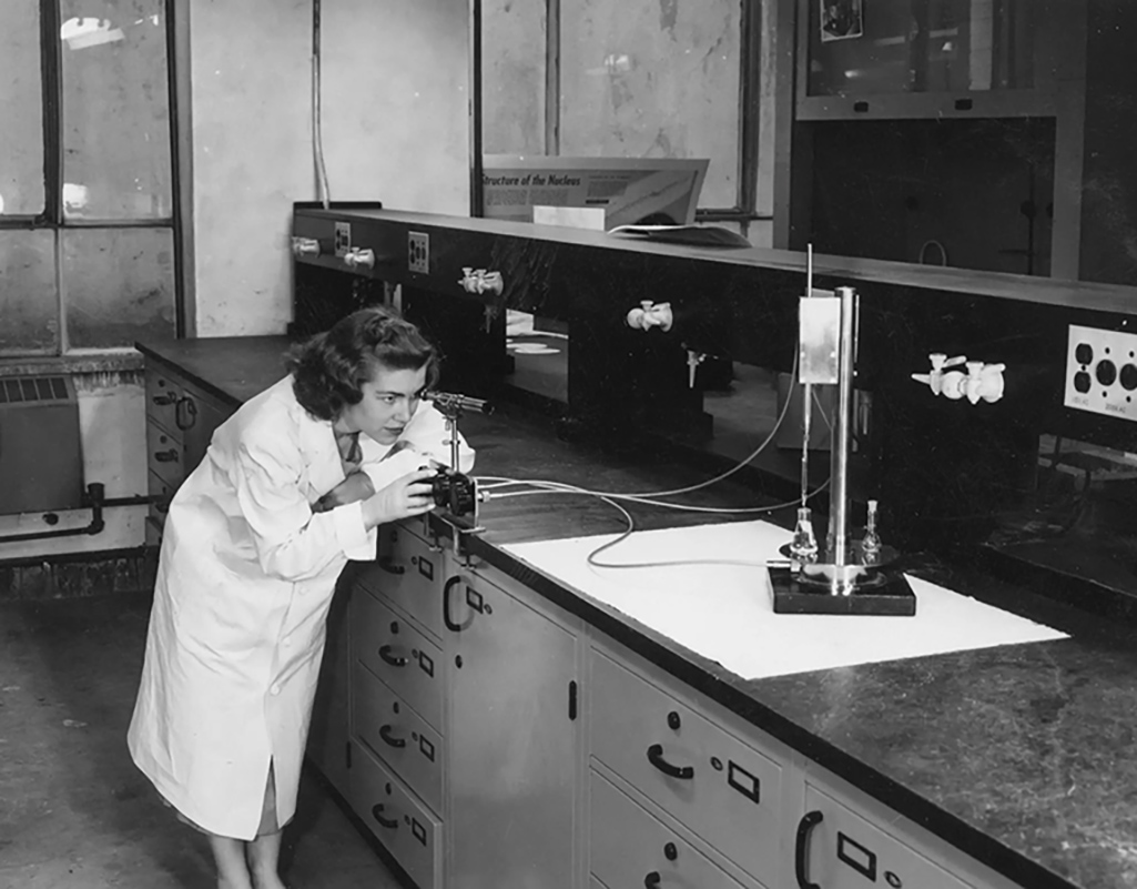 Black and white photo of a person wearing a lab coat peering through scientific equipment.