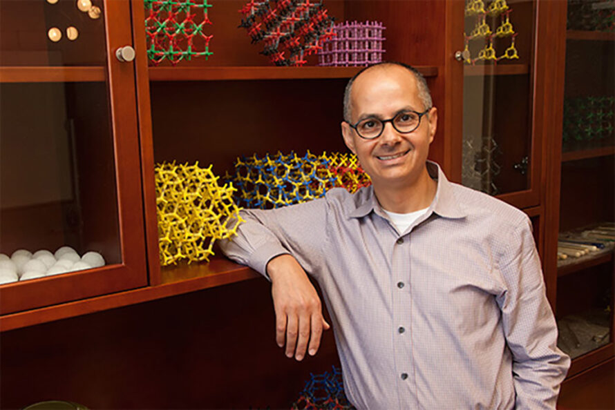 Person wearing glasses and long sleeve shirt leaning against wooden shelves holding chemistry models