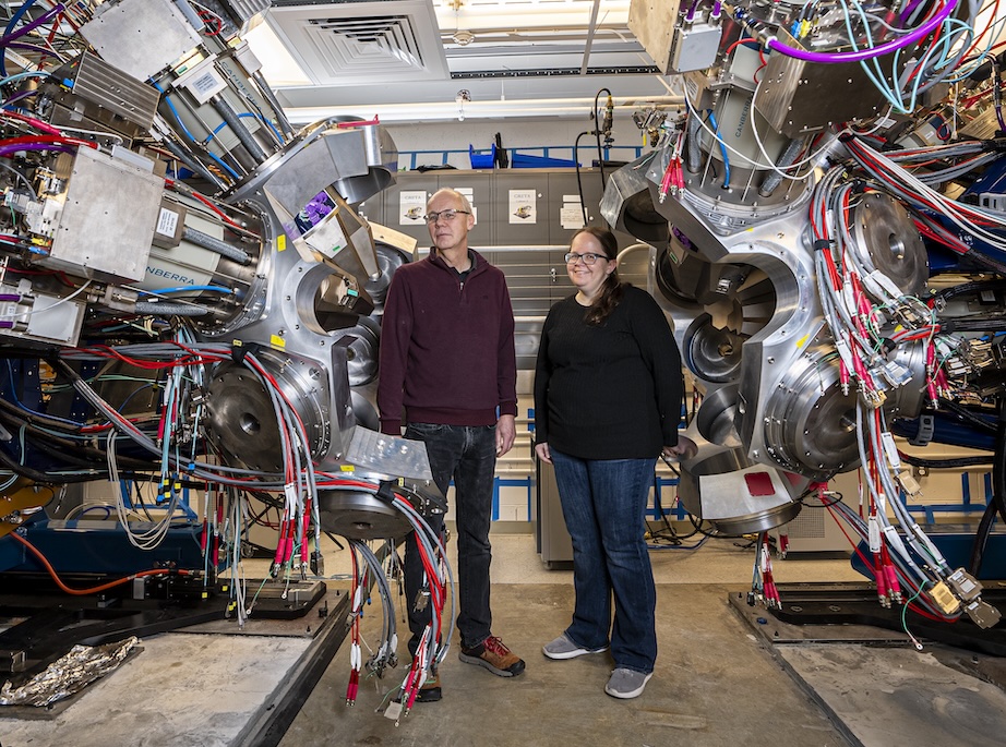 Two researchers stand between large, complex pieces of scientific equipment filled with cables, sensors, and metal components inside a laboratory setting.
