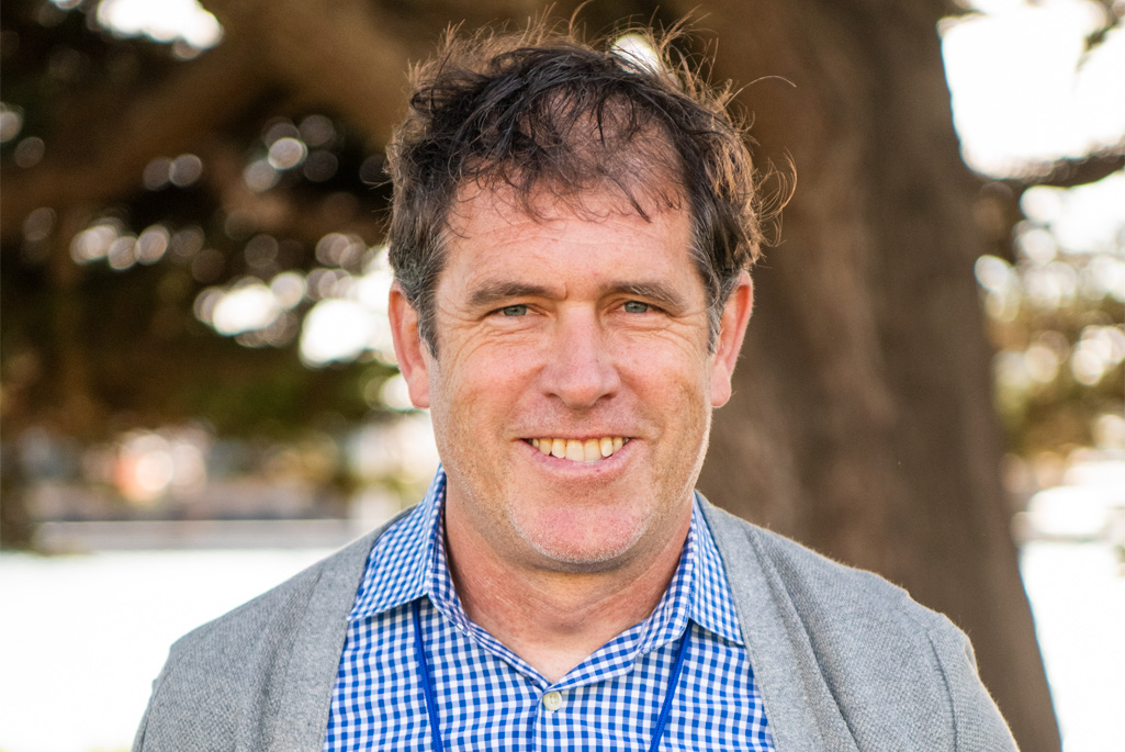 Dula Parkinson, a person with short brown hair wearing a grey jacket over a blue and white collared checkered shirt, smiling.