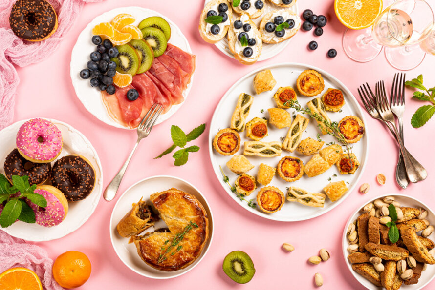Top view flat lay of holiday dinner party table, with assorted food and drinks on pink background.