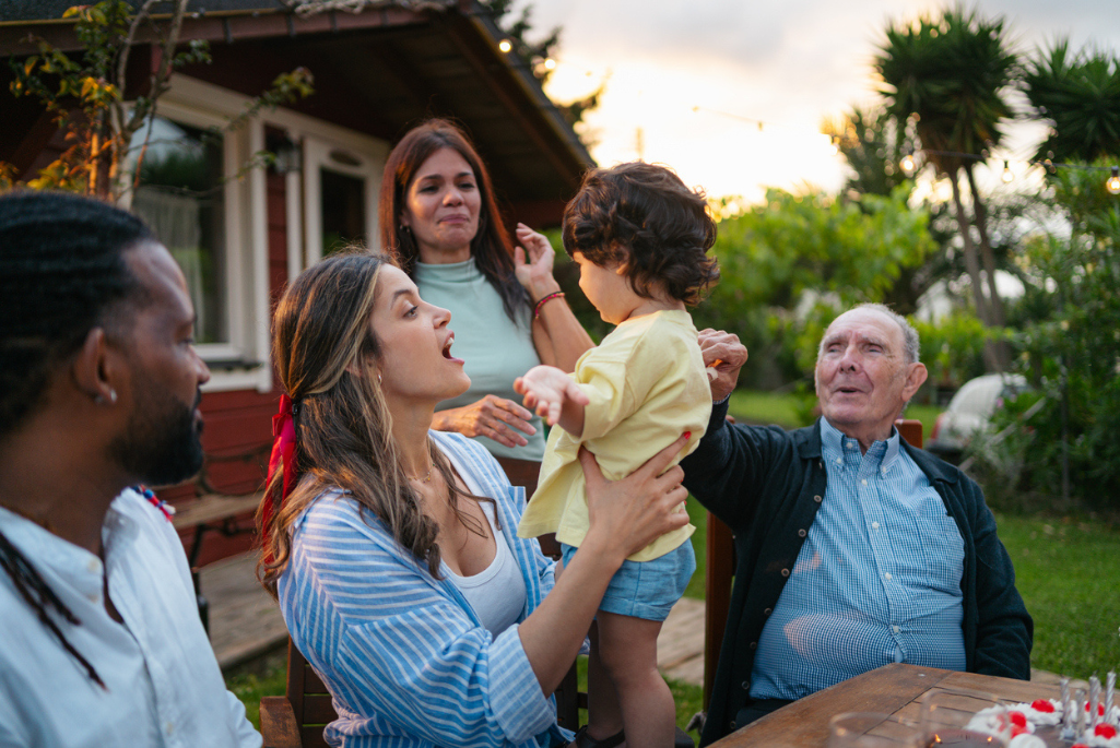 Photo of multi-generational family, including a toddler, parents, and grandparents, gathers for a joyful celebration in their backyard
