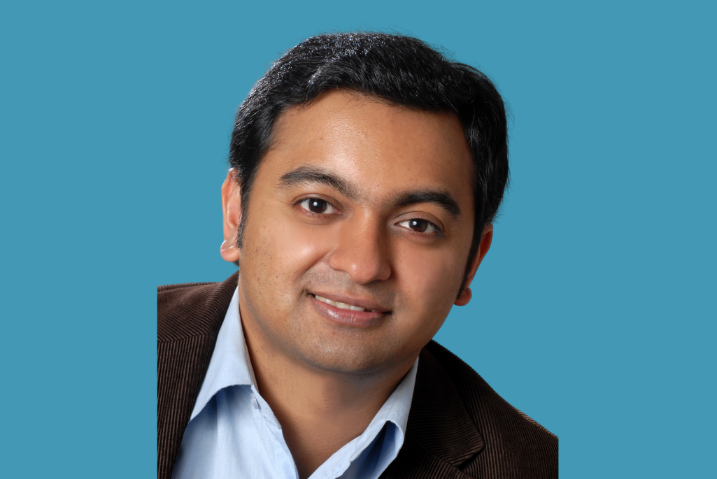 Headshot of person with short black hair wearing a brown jacket over blue shirt and smiling.