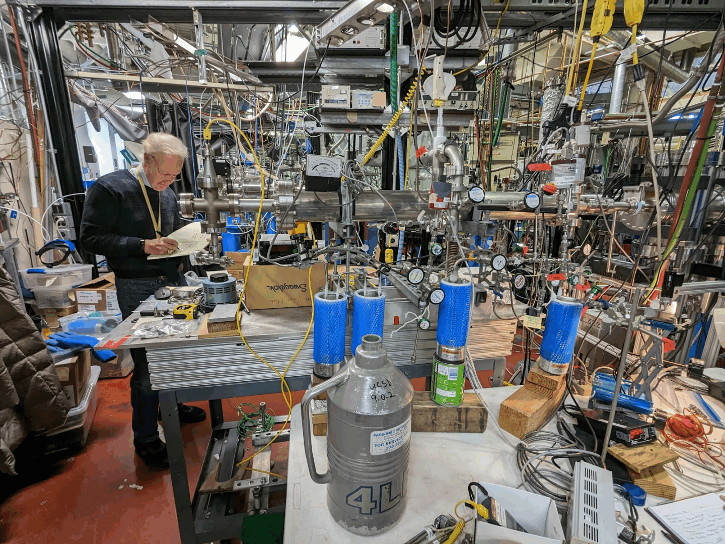 A researcher stands at a crowded laboratory bench filled with metal tubing, pressure gauges, blue filter canisters, and electrical wiring.