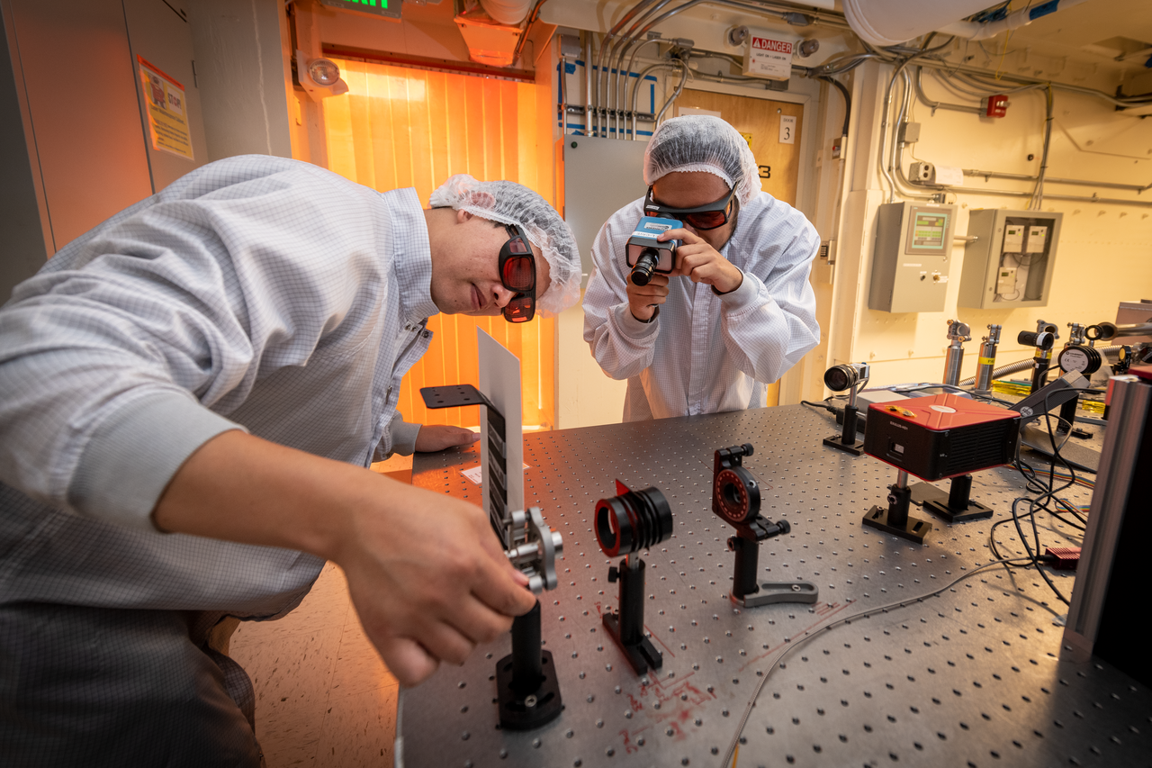 Two scientists in a lab wearing safety equipment and examining a sheet fixed in place between black clamps
