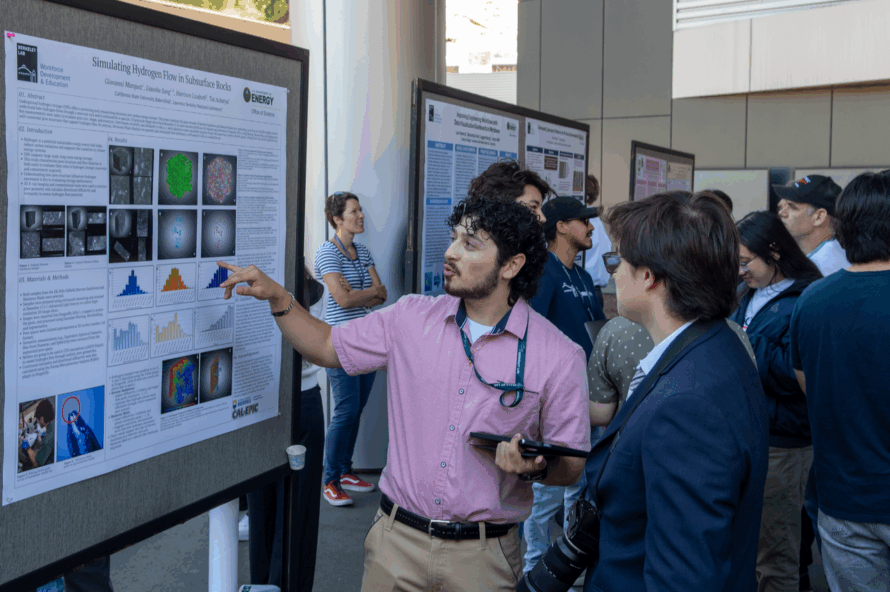 Person wearing pink shirt and tan pants points toward mounted science poster with another person looking on