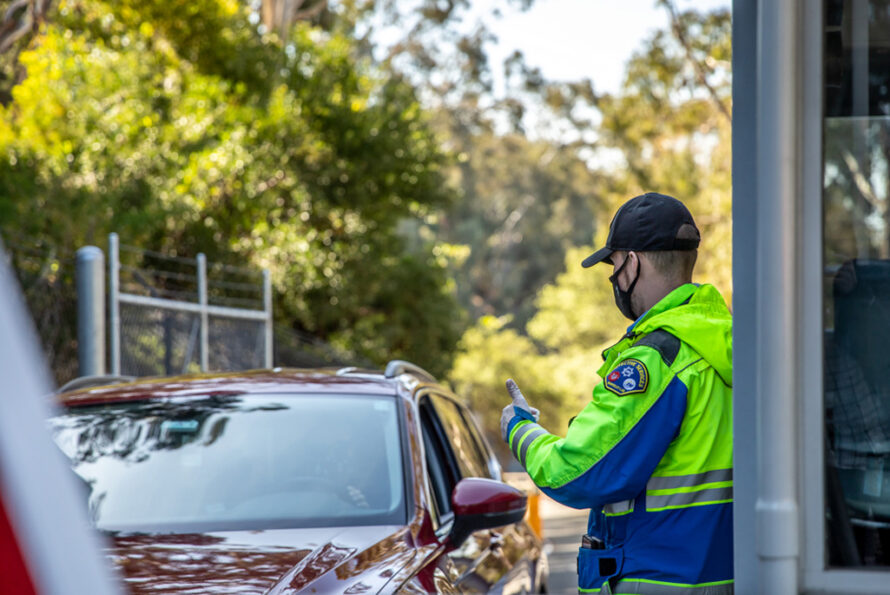 A security guard in a neon safety jacket and a hat gives a thumbs up to the driver of a car entering through the Lab's secured gate.