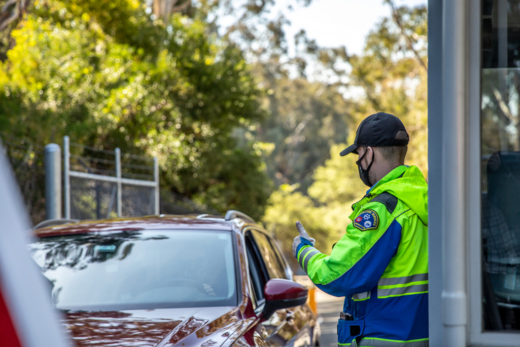 A security guard in a neon safety jacket and a hat gives a thumbs up to the driver of a car entering through the Lab's secured gate.