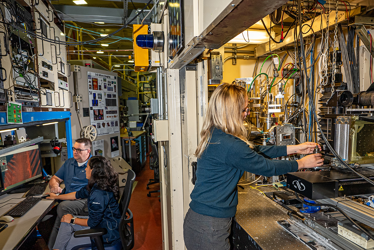 Two researchers in the background of a beamline laboratory look toward a computer with colorful microtomography imagery. A researcher is in the foreground making adjustments to an experimental setup.