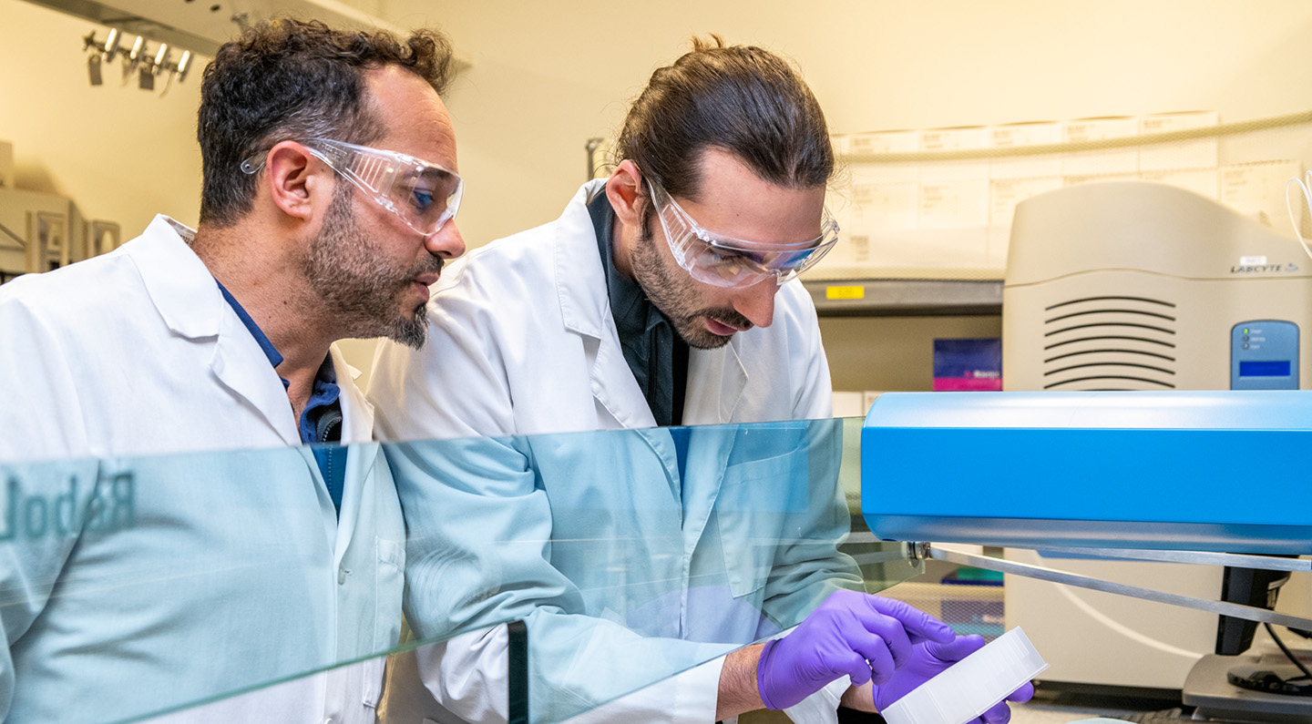 Two researchers in a lab space wear personal protective equipment and look down towards a sample.