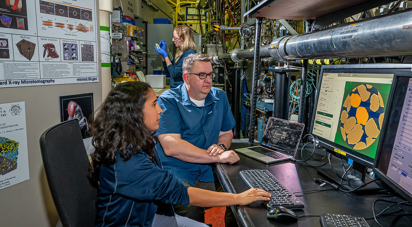Two researchers in the foreground of a beamline laboratory look toward a computer with colorful microtomography imagery. A researcher is in the background examining an experimental setup.