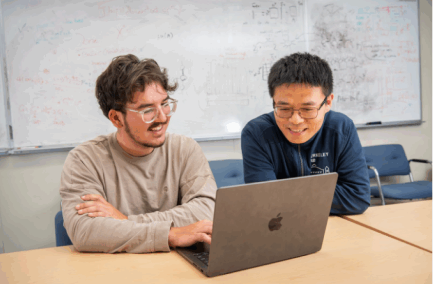 Two persons sitting at table, one typing on laptop.