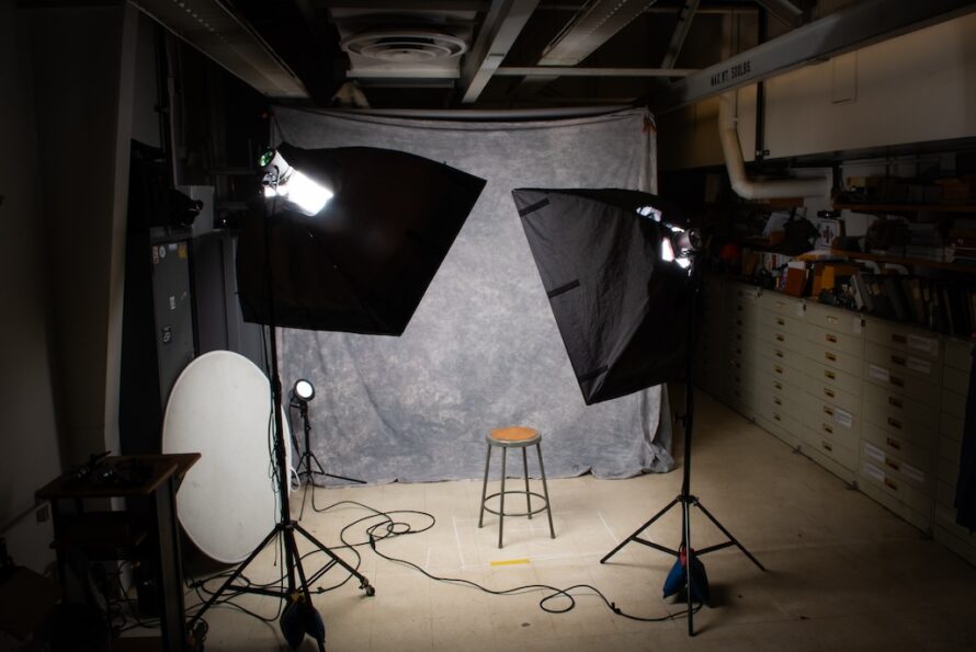 Photo studio scene with brightly lit space with wooden stool in the center against a gray background with tripod photography equipment in the foreground.