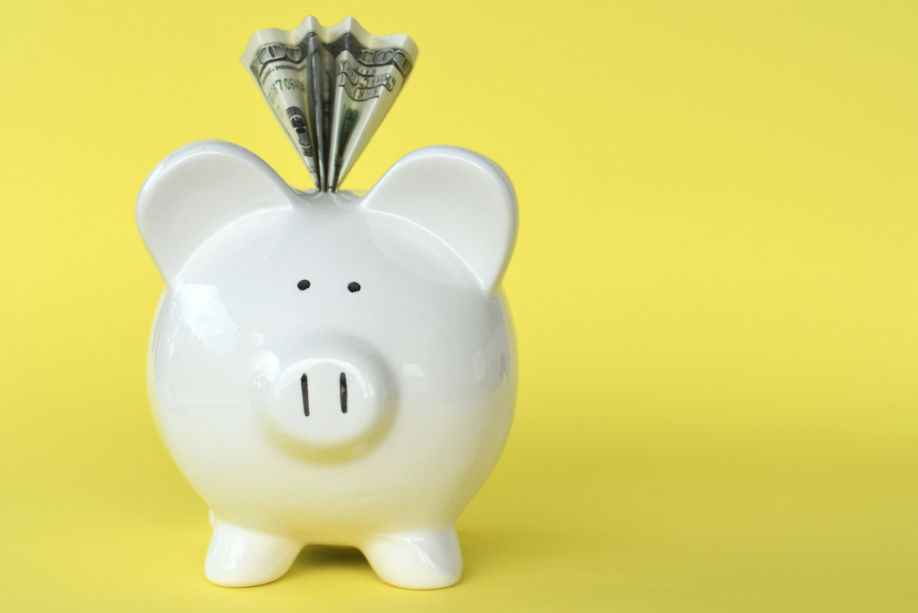 white ceramic piggy bank with dollar coming out of the top, against a yellow background