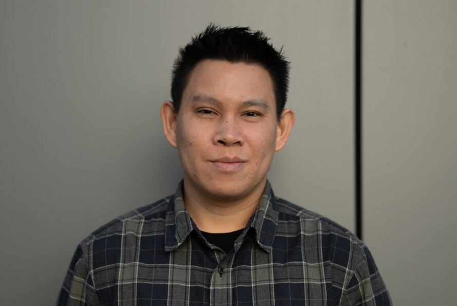 Portrait of Jimmy Vo, a person with short dark hair wearing a plaid flannel shirt, smiling for a portrait against a wall.