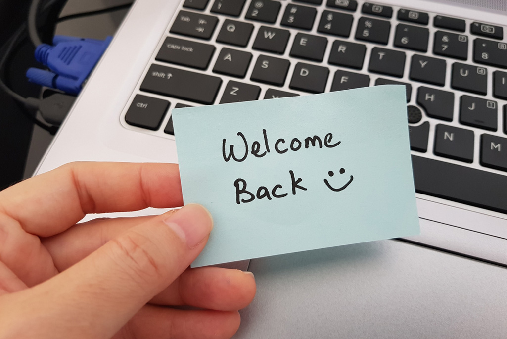 Hand holding a blue note that says, "Welcome Back" in front of a laptop.