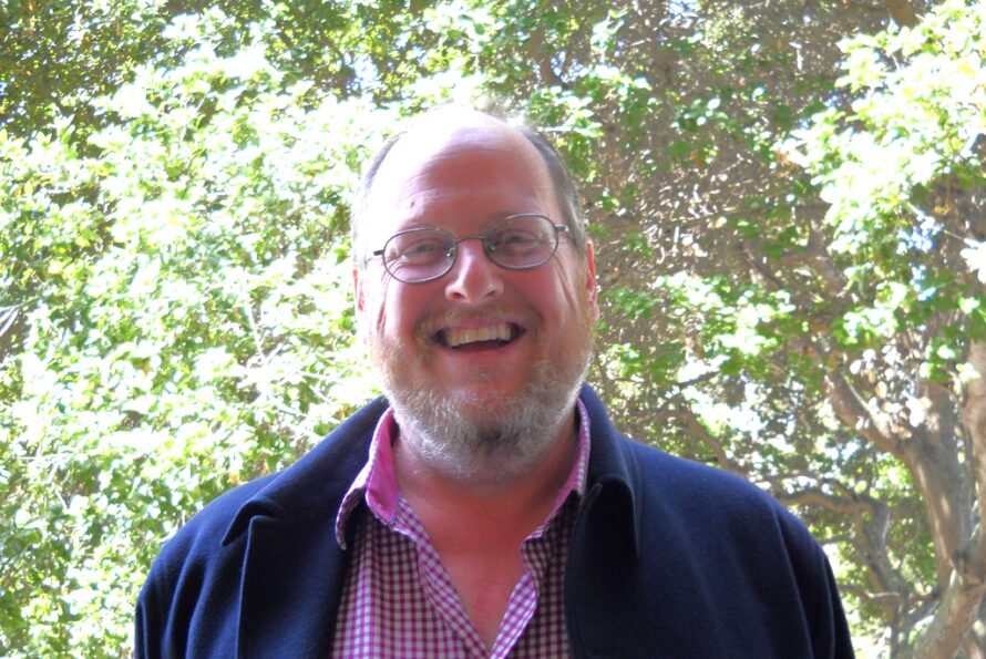 Headshot of smiling person with gray beard and wire glasses wearing a red shirt and blue sweater