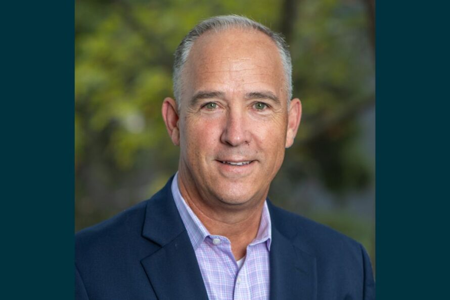 Person with short gray hair wearing a navy blazer over a purple checkered collared shirt, smiling for a portrait with greenery in the background.