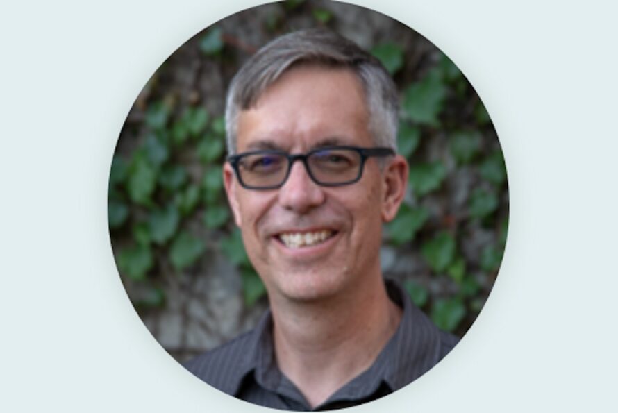 Circular headshot of a smiling person with short dark hair and black glasses wearing a gray shirt