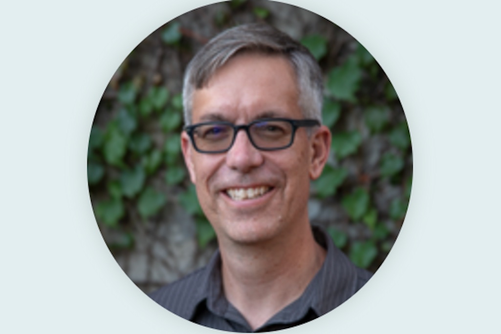 Circular headshot of a smiling person with short dark hair and black glasses wearing a gray shirt