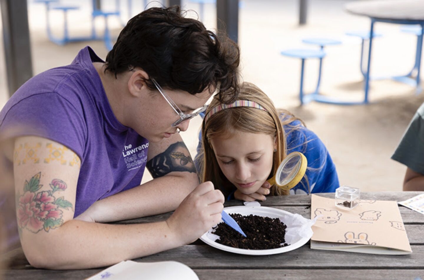 Two persons seated at wood table: one person with short dark hair, colorful tattoos on both arms probes a mound of brown crystals while other person with long blond hair look on