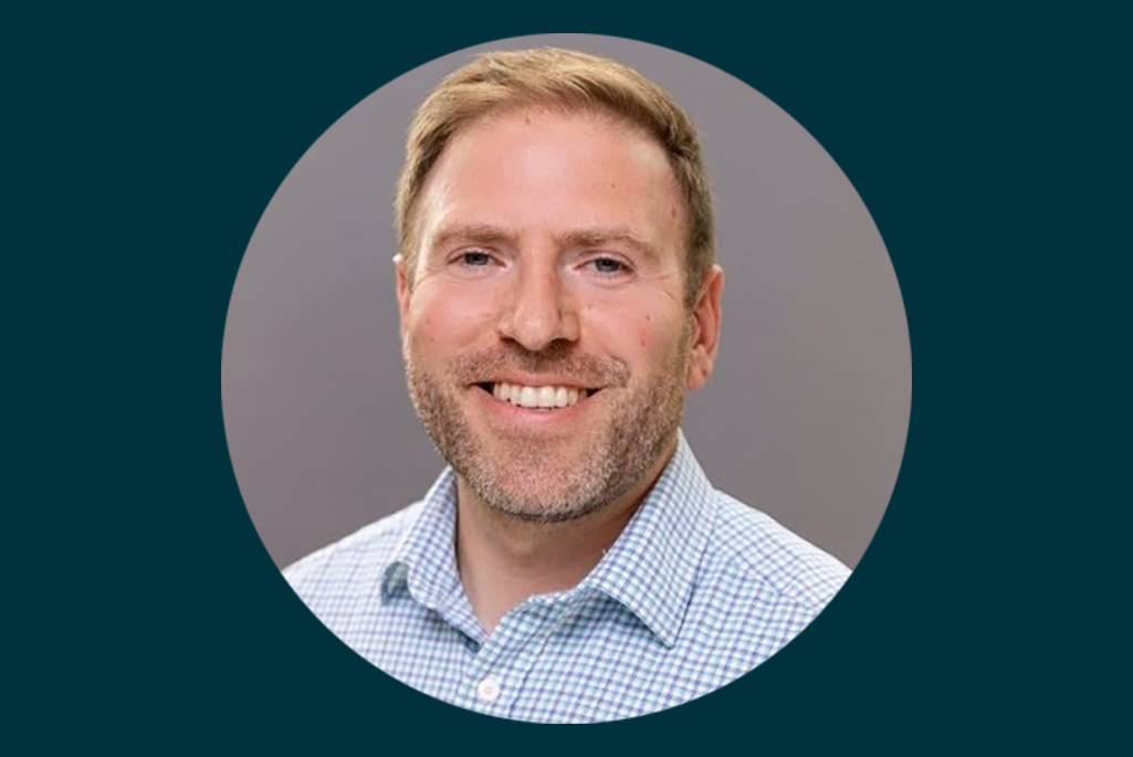 Headshot of person wearing blue and white checked shirt with red hair and beard smiling