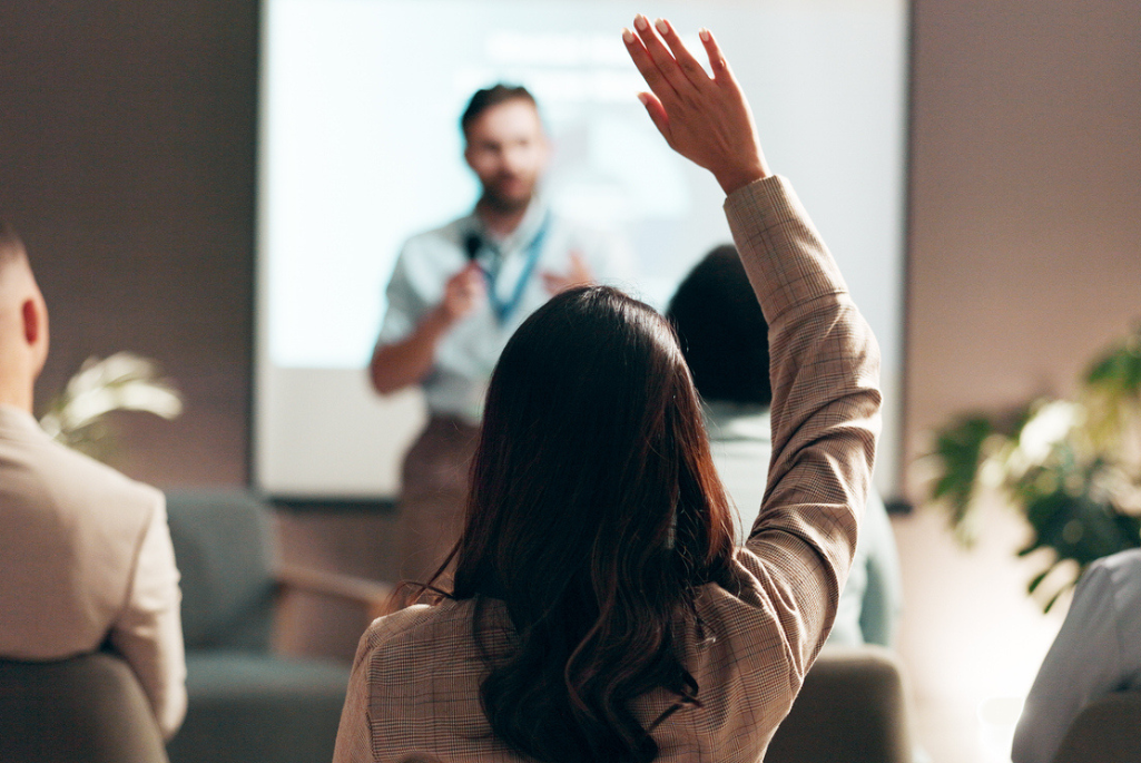 A person in a classroom setting holds up their hand in the foreground with the instructor visible standing in the background