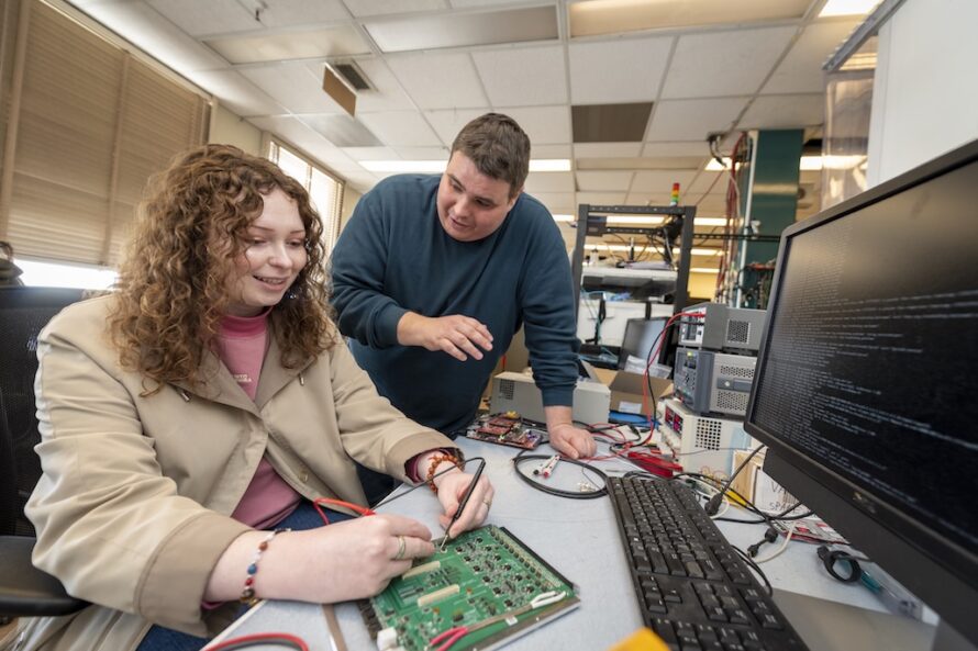 Seated person with long brown hair wearing tan jacket manipulating device on a desktop while a standing person with short dark hair in a green sweater looks on