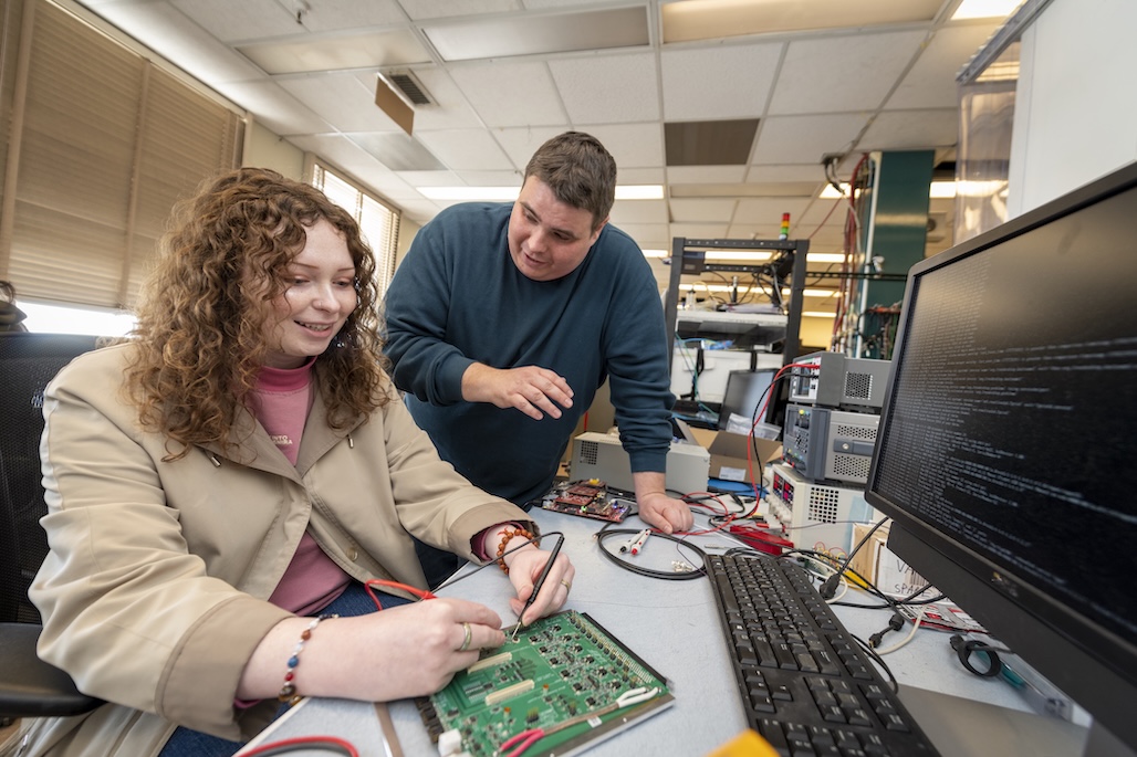 Seated person with long brown hair wearing tan jacket manipulating device on a desktop while a standing person with short dark hair in a green sweater looks on