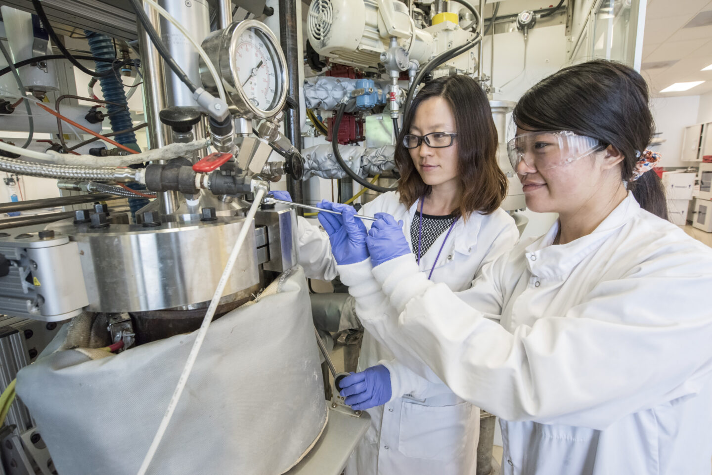 Two scientists with long dark hair, wearing glasses and protection gloves, manipulate device