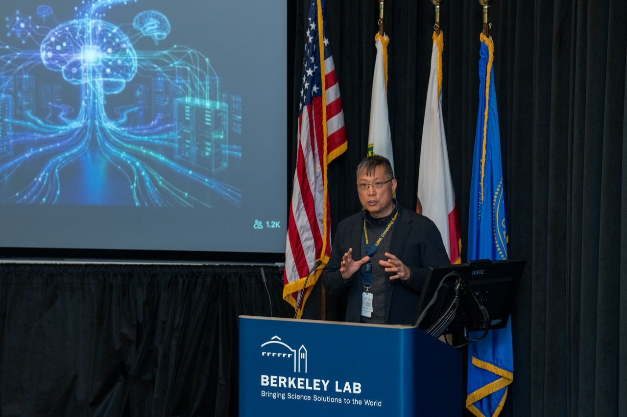 Person standing behind podium gesturing with their hands with four flags and a wall-mounted monitor in the background