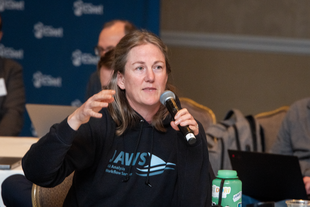 Person with long blonde hair wearing black hoodie sweatshirt and holding a microphone in one hand and gesturing with the other hand.