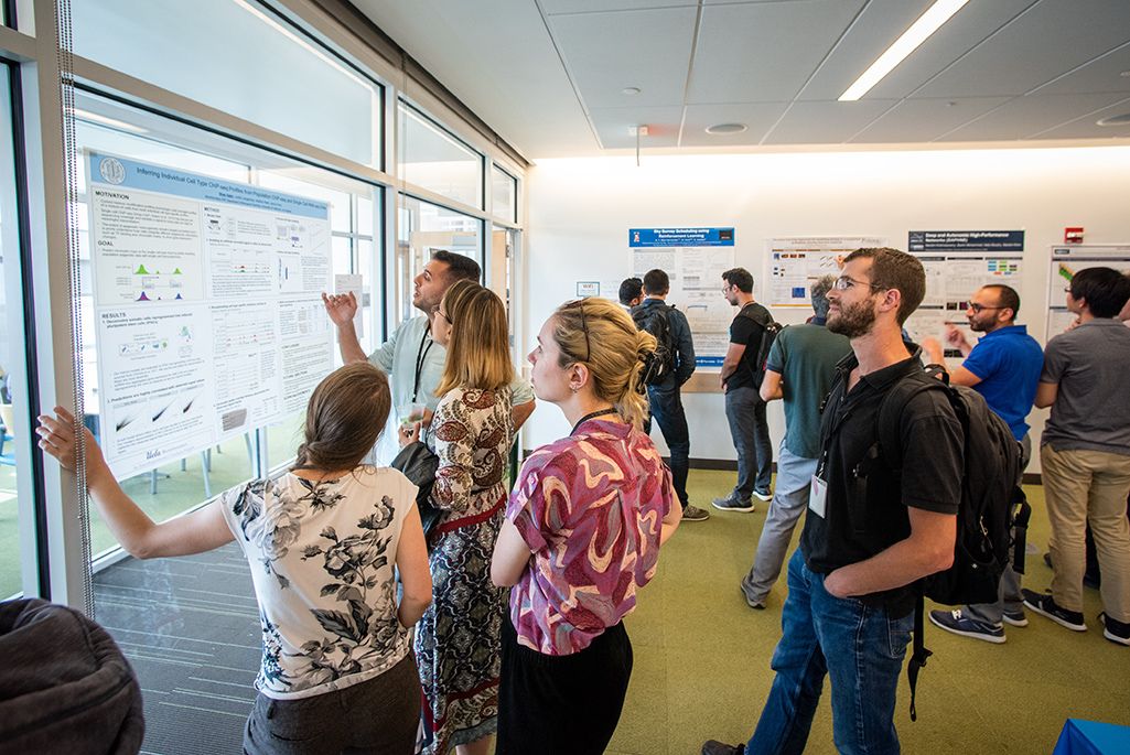 Group of persons standing in front of wall-mounted poster board with one person gesturing toward the board